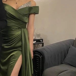 Grön offshoulder klänning med glittrig detalj - Elegant grön klänning med offshoulder-design och glittrig detalj vid bysten. Klänningen har en smickrande drapering och en hög slits som ger en sofistikerad look.