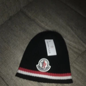 Svart mössa från Moncler - Snygg svart mössa från Moncler med röd och vit randdetalj. Den har en broderad logotyp framtill och är perfekt för kyliga dagar. Skriv för pris !!