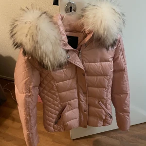 Rosa dunjacka från Rockandblue - Säljer en snygg rosa dunjacka från Rockandblue med fluffig päls på huvan. Jackan har dragkedja och knappar framtill samt två praktiska fickor med dragkedja. Perfekt för kyliga dagar!