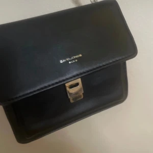 Svart axelväska från David Jones - Snygg svart axelväska från Savlone med en elegant kedjerem och stilrent spänne. Perfekt för att ge en touch av klass till din outfit. Väskan har en kompakt design som gör den idealisk för att bära det viktigaste.