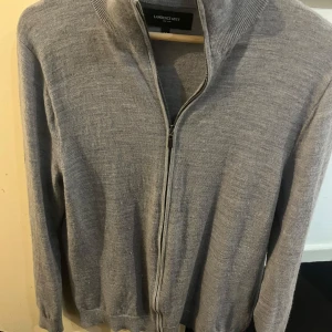 Grå merinoulltröja från Lawrence Grey - Säljer en stilren grå tröja från Lawrence Grey i 100% merinoull. Tröjan har en dragkedja framtill och ribbade detaljer vid krage och ärmslut. Perfekt för kyligare dagar.