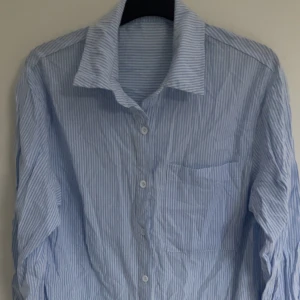 Blå- och vitrandig skjorta - Snygg blå- och vitrandig skjorta med klassisk krage och knappar framtill. Skjortan har en bröstficka och långa ärmar, perfekt för en stilren look.