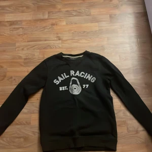Svart tröja från Sail Racing - Snygg svart tröja från Sail Racing med tryck framtill. Tröjan har långa ärmar och rund halsringning. Perfekt för en avslappnad stil.
