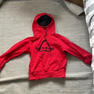 Röd hoodie från SOS - Säljer en cool röd hoodie från SOS med svart tryck på framsidan. Den har en justerbar huva och en stor ficka framtill. Perfekt för en avslappnad stil.