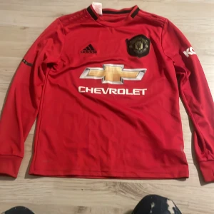 Röd långärmad fotbollströja från Adidas - Säljer en röd långärmad fotbollströja från Adidas med svart krage och klubbmärke på bröstet. Tröjan har Chevrolet-logga i guld och vitt framtill. Perfekt för fotbollsfans!