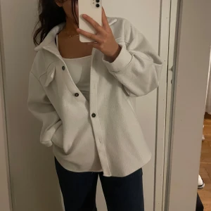 Vit overshirt med knappar - Snygg vit overshirt med knappar framtill och en bröstficka. Perfekt för lager-på-lager-stil och passar till både jeans och kjol. Enkel och stilren design som funkar till många tillfällen.