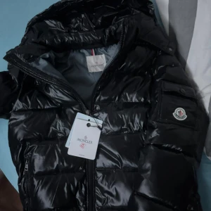 Moncler maya - Snygg svart pufferjacka från Moncler med glansig finish och dragkedja framtill. Jackan har en huva och två praktiska fickor med dragkedja. Perfekt för kyliga dagar. Nfc och allt funkar 