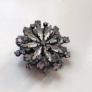 Glittrig brosch med kristaller - Elegant brosch i form av en blomma, dekorerad med gnistrande kristaller i silverfärg. Perfekt för att ge en extra touch av glamour till din outfit. Broschen har en säkerhetsnål på baksidan för enkel fastsättning.