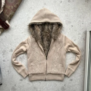 Söt tröja med päls - Mysig beige stickad hoodie med fuskpäls i luvan och dragkedja framtill. Den har ett dekorativt stickat mönster och långa ärmar med ribbade muddar. Perfekt för kyliga dagar.