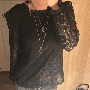 Svart spetsblus med volangdetaljer - Elegant svart spetsblus med volangdetaljer på axlarna. Blusen har långa ärmar och en fin knytning vid halsen. Perfekt för en stilfull look.