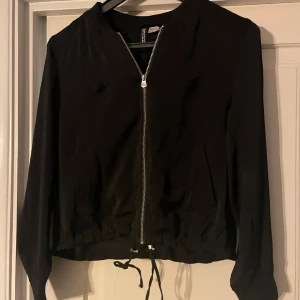 Svart tunn jacka/tröja från H&M - Snygg tunn svart jacka/tröja från H&M:s Divided-kollektion. Jackan har en dragkedja framtill och långa ärmar. Perfekt för en stilren look.
