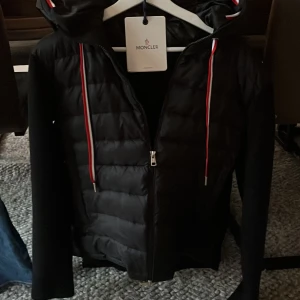 Svart jacka från Moncler - Snygg svart jacka från Moncler med quiltad design och dragkedja. Jackan har en huva med färgglada snören och Moncler-logga på ärmen. Perfekt för kyligare dagar. 100% äkta 