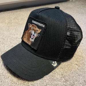 Svart keps från The Farm - Snygg svart keps från The Farm med ett broderat sabertandmotiv på framsidan. Kepsen har ett justerbart spänne bak och meshpaneler för extra ventilation.