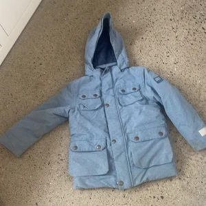 Blå vinterjacka från Molo - Säljer en snygg och praktisk blå vinterjacka från Molo. Jackan har en luva och är utrustad med både dragkedja och knappar för extra skydd mot kylan. Den har flera fickor med knappar, perfekt för att förvara småsaker. Perfekt för kalla vinterdagar! För barn 122 cm 