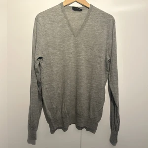Grå ulltröja från Ralph Lauren - Säljer en stilren grå tröja från Ralph Lauren i 100% merinoull. Den har en v-ringad design och långa ärmar, perfekt för både höst och vinter. Tröjan är i bra skick men har ett litet märke på framsidan. Supermjuk och bekväm att ha på sig!