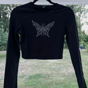 Croppad långärmad tröja - En croppad svart långärmad tröja med strass-detalj i form av en fjäril på bröstet 🦋 En aning liten i storleken då jag normalt bär S. Använd max 5 gånger 💙  Längd: 42 cm Axel: 35 cm Byst: 80 cm