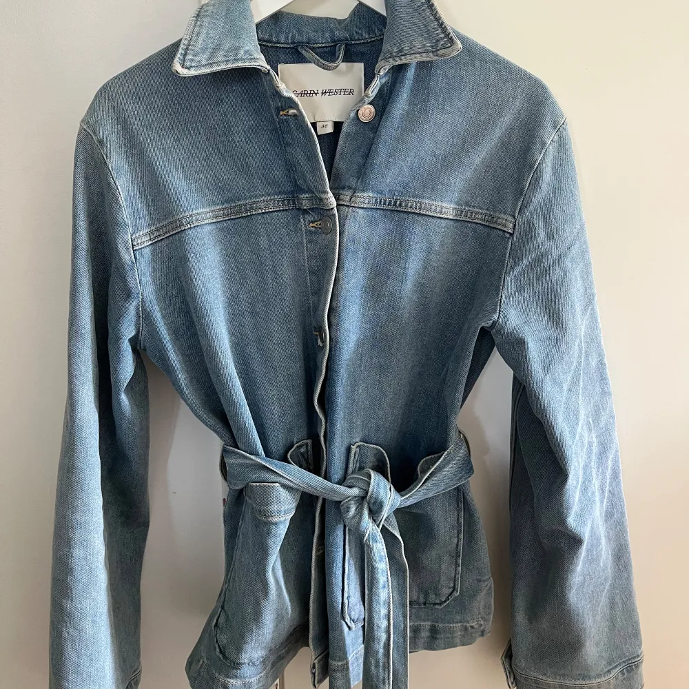 Säljer en snygg jeansjacka från Carin Wester i storlek 36. Jackan har en klassisk blå denimfärg och är utrustad med ett bälte i midjan för en mer figurnära passform. Den har långa ärmar och stora fickor framtill, perfekt för en avslappnad stil. Passar perfekt till vår och höst!. Jackor.