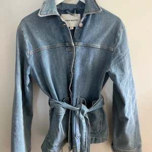 Jeansjacka från Carin Wester - Säljer en snygg jeansjacka från Carin Wester i storlek 36. Jackan har en klassisk blå denimfärg och är utrustad med ett bälte i midjan för en mer figurnära passform. Den har långa ärmar och stora fickor framtill, perfekt för en avslappnad stil. Passar perfekt till vår och höst!