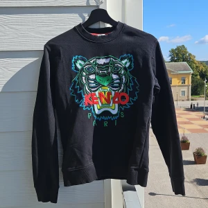 Svart Kenzo tröja med tigerbroderi - Säljer en supercool svart tröja från Kenzo med ett stort, färgglatt tigerbroderi på framsidan. Tröjan har långa ärmar och är i storlek XS. Perfekt för att ge din outfit en edgy touch! 🐯