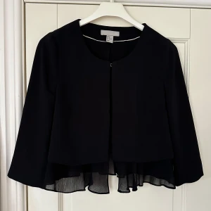 Kort blazer med detaljer - Svart kortare blazer med detaljer nedtill och kortare ärm från H&M i storlek 38 (ej full längd på varken blazern eller ärmarna). En hake framtill för att kunna stänga, perfekt över en klänning. Knappt använd så i gott skick.