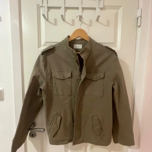 Olivgrön jacka från Neo Noir - Säljer en snygg olivgrön jacka från Neo Noir. Den har en stilren design med långa ärmar och praktiska fickor framtill. Jackan är perfekt för höst och vår, och har en bekväm passform. Den stängs med knappar och har en klassisk krage.