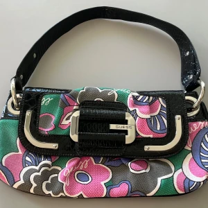 Guess handväska oanvänd utan tag. - Guess handväska i flerfärgat tyg med ”guess” tryckt  på. Läderliknande handtag och på kant runt lock. Silvertonad hårdvara med ”Guess” präglat. Invändig blixtlåsficka och en extra liten ficka  inuti. Silkesliknande tyg på insidan. Magnetisk stängning
