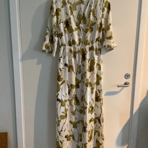 Vit och grön långklänning med blommigt mönster - Säljer en superfin långklänning i vitt med grönt blommigt mönster. Klänningen har en v-ringad design och trekvartsärmar, vilket gör den perfekt för både vardag och fest. Materialet känns lätt och luftigt, perfekt för varmare dagar.