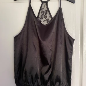 Svart linne från XOXO Friends - Säljer ett snyggt svart linne från XOXO Friends. Det har en elegant spetsdetalj på ryggen och är gjort i ett glansigt material som ger en lyxig känsla. Perfekt för en utekväll eller fest!