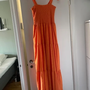 Orange maxiklänning - En orange klänning som man knyter vid axlarna. Klänningen är i en A-linjeform och har en elastisk midja. Storlek 14 eller 40. Mycket stretchig upptill