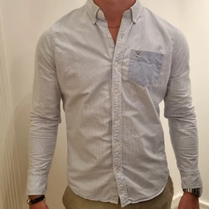 Ljusblå randig skjorta  - Ljusblå randig skjorta av 100% bomull, med fina detaljer. Passar för både formella och vardagliga tillfällen och går smidigt att kombinera med andra plagg. Väldigt bra skick, inköpt 2011 när Hollister öppna sin första butik i Sverige. 