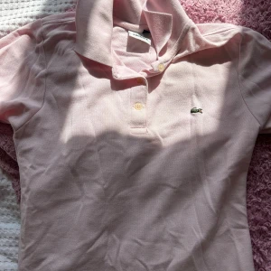 Ljusrosa pikétröja från Lacoste - Säljer en snygg ljusrosa pikétröja från Lacoste. Den har en klassisk krage och två knappar framtill. Perfekt för en avslappnad men stilren look. Tröjan är i bra skick och redo för nya äventyr!