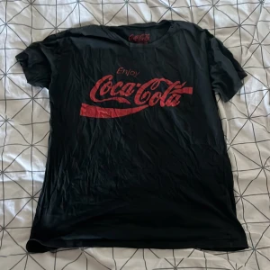 Svart Coca-Cola t-shirt från Pull&Bear - Säljer en svart t-shirt från Pull&Bear med det klassiska Coca-Cola-trycket i rött på framsidan. T-shirten är kortärmad och i bomull, perfekt för en avslappnad stil. Den är i storlek S och har en normal passform. Perfekt för alla Coca-Cola-fans! 🥤