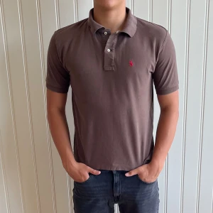 Brun pikétröja från Ralph Lauren - Säljer en snygg brun pikétröja från Ralph Lauren. Den har en klassisk krage med knappar och en liten röd broderad logga på bröstet. Perfekt för en stilren och avslappnad look! 180cm 75kg