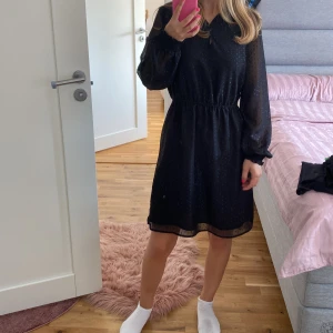 Svart klänning från Camaïeu - Säljer en elegant svart klänning från Camaïeu. Den har ett diskret prickigt mönster och långa ärmar med smockdetaljer vid axlarna. Klänningen har en knappslå framtill och en elastisk midja för en bekväm passform. Perfekt för både vardag och fest!