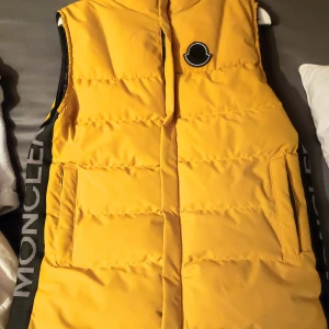 Gul dunväst från Moncler - Säljer en snygg gul dunväst från Moncler i mycket bra skick. Västen har en dragkedja framtill och stora fickor. Perfekt för höst och vår, och den har en cool logga på bröstet. Insidan är svart med ett unikt mönster. Passar perfekt för en casual look eller en dag ute i stan.