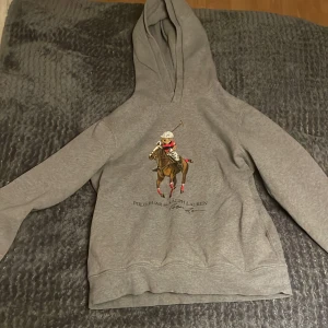 Grå hoodie från Polo Ralph Lauren - Säljer en snygg grå hoodie från Polo Ralph Lauren med en cool polobjörn på framsidan. Perfekt för en avslappnad stil och passar bra till både jeans och joggers. Hoodien är i bra skick och superbekväm att ha på sig. lite av trycket har dock gått bort. storlek 14-16 och passar mig som är runt 168cm lång. finns inte att köpa längre!