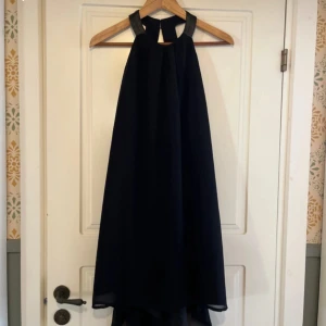 Svart axelbandslös klänning - Snygg svart axelbandslös klänning i ett elegant och flytande material. Perfekt för festliga tillfällen. Klänningen har en stilren design med en halterneck-stil och en luftig passform.