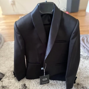 Barn kavaj  - Barnkostym som aldrig använts. Helt ny med lappen på. Märket är från Turkiet och i lilla påsen finns en fluga och ett till tillbehör. Hela kitet köptes för 3600 kr som ny men vid snabb affär kan man diskutera priset. Passar till barn i två års åldern. Kostym + kostymbyxor. Köparen står för frakten annars går det bra att hämta upp den 