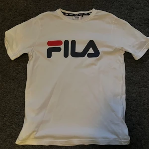 Vit t-shirt från Fila - Säljer en snygg vit t-shirt från Fila. Den har en stor logga i mörkblått och rött på bröstet. Perfekt för en casual look eller träning. T-shirten är kortärmad och har en normal passform. Använd ett fåtal gånger men ser ut som ny. Storlek 158/164 (13-14 år)