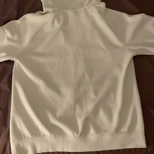 Vit hoodie från Nike - Säljer en vit hoodie från Nike i bra skick. Den har en dragkedja framtill och en justerbar huva med svarta snören. Perfekt för både vardag och träning. Det finns en liten fläck på framsidan, men annars är den i fint skick. Jag är 183cm och pris kan sänkas