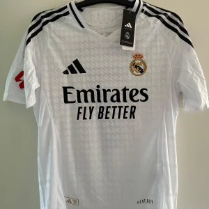 Vit Real Madrid fotbollströja från Adidas Vini JR - Säljer en vit Real Madrid fotbollströja från Adidas i storlek L. Tröjan är i nytt skick med korta ärmar och har klubbens emblem på bröstet. Den är tillverkad i ett lätt och andningsbart material med 'HEAT.RDY' teknologi för optimal komfort. Perfekt för alla Real Madrid-fans! Storlek L