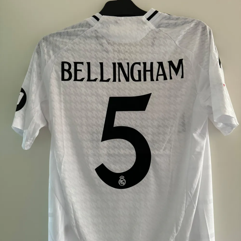 Säljer en vit Real Madrid fotbollströja från Adidas i nytt skick. Tröjan har korta ärmar och är prydd med klubbens emblem och sponsorlogga. Baksidan har namnet 'Bellingham' och nummer 5. Storlek L. T-shirts.