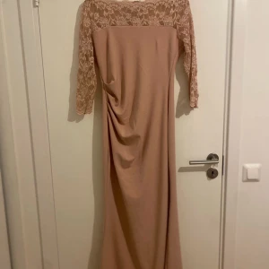 Beige långklänning från Goddiva - Elegant beige långklänning från Goddiva med vackra spetsdetaljer på överdelen och trekvartsärmar. Perfekt för festliga tillfällen med sin stilrena design och mjuka fall. Klänningen har en draperad detalj framtill som ger en smickrande passform.