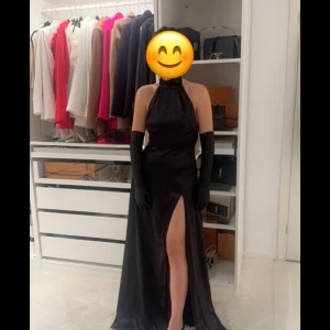 Svart långklänning med vit rosett - Elegant svart långklänning med en djärv slits och en stor vit rosett i ryggen. Klänningen är axelbandslös och har en öppen rygg, vilket ger en sofistikerad look. Perfekt för festliga tillfällen!