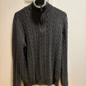 Tröja - Säljer en snygg mörkgrå stickad tröja med dragkedja vid halsen. Perfekt för kyliga dagar med sitt kabelstickade mönster och ribbade detaljer vid krage, ärmslut och nederkant. Passar både till vardags och lite finare tillfällen. Supermysig och stilren!