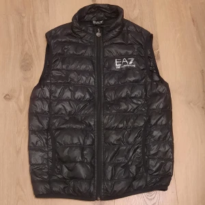 Svart dunväst från Emporio Armani - Säljer en svart dunväst från Emporio Armani EA7. Västen har en dragkedja och är quiltad för extra värme. Den är i bra skick men har en liten skada på insidan. Perfekt för höst och vår! Den är storlek S men passar även XS 🙌