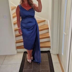 Blå sidenklänning - Elegant blå sidenklänning med omlott-design och en vacker drapering. Klänningen har en asymmetrisk längd och är ärmlös, perfekt för festliga tillfällen. Den djupa blå färgen ger en lyxig känsla.