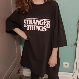 Svart Stranger Things t-shirt från H&M Divided - Säljer en svart oversized t-shirt från H&M Divided med Stranger Things-tryck i vitt. Perfekt för alla fans av serien! T-shirten har trekvartsärmar och är i bomull, vilket gör den superbekväm. Passar perfekt till en avslappnad stil eller som en del av en cool outfit.