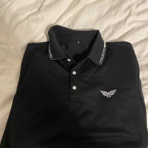 Svart pikétröja med logga - Säljer en stilren svart pikétröja med en snygg logga på bröstet. Tröjan har en klassisk krage med textdetaljer och knappar framtill. Perfekt för både vardag och fest! 🖤