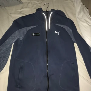 Mörkblå jacka från Puma - Säljer en snygg mörkblå jacka från Puma i samarbete med Mercedes. Jackan har en dragkedja framtill och är långärmad med ett coolt mönster på ärmarna. Perfekt för höst och vår! 🏎️